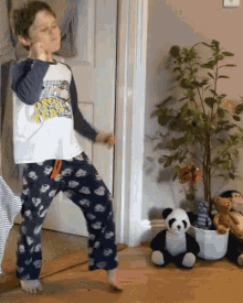 a young boy in pajamas is dancing in front of a plant and stuffed panda bear