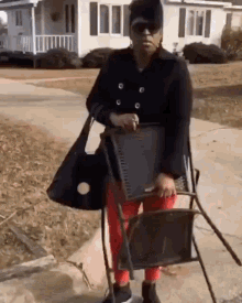 a woman in a black coat and red pants is holding a chair and a purse