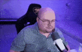 a bald man wearing glasses and a plaid shirt sitting in front of a microphone