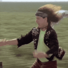 a man with long hair wearing a headband and a jacket is dancing in a field .