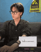a man wearing glasses sits at a desk with a sign that says i add a comment