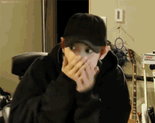 a man wearing a baseball cap is covering his mouth with his hands while sitting in front of a guitar .