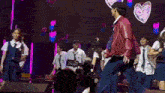 a group of young people are dancing on a stage with hearts on the wall behind them .