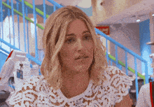 a blonde woman wearing a white lace top looks at the camera