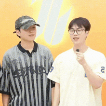 two young men are standing next to each other in front of an orange wall . one of the men is wearing a hat and glasses .