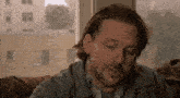 a man with long hair and a beard is sitting on a couch in front of a window with his eyes closed .