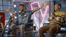 two men are sitting in chairs with their arms outstretched in front of a neon sign that says nbc