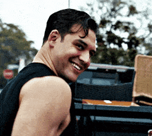 a man in a black tank top is smiling while standing next to a truck .