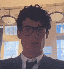 a man with curly hair wearing glasses and a suit looks at the camera