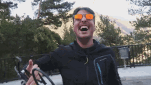 a man wearing sunglasses and a black jacket laughs with his mouth open