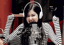 a woman wearing headphones and a striped shirt is standing in front of a microphone and smiling .