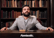 a man in a suit is sitting at a desk in front of a bookshelf and says fantastic .