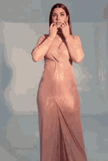 a woman in a long pink dress is standing in a room and touching her face .