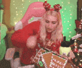 a woman in a red sweater is holding a gingerbread house in front of a christmas tree .