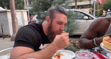 a man with a beard is eating a slice of pizza while wearing a black shirt with the letter m on it