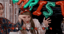 two girls are sitting at a table with balloons in the background and one girl is wearing a christmas headband .