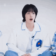 a young man wearing a white shirt with a blue flower on the sleeve is sitting down with his mouth open .