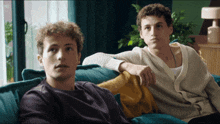 two young men are sitting on a blue couch and one of them is wearing a white cardigan