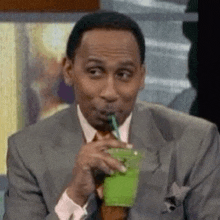 a man in a suit and tie is drinking a green smoothie from a plastic cup with a straw .