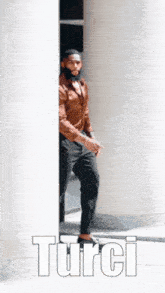 a man in a brown shirt and black pants is standing behind a white pillar and the word turci is on the bottom