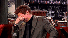 a man in a suit and tie is sitting on a couch covering his face with his hand .