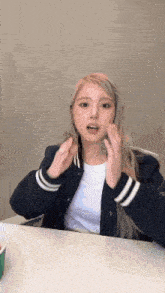 a woman is sitting at a table with her hands on her face . she is wearing a baseball jacket and a white shirt .