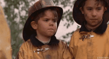 a boy wearing a fireman 's helmet stands next to another boy wearing a fireman 's jacket