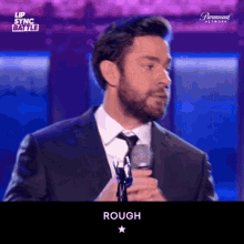 a man in a suit and tie is holding a microphone and the word rough is on the screen