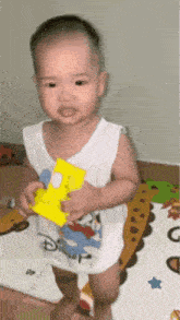 a baby in a white tank top is standing on a rug holding a toy .