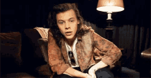 harry styles is sitting on a couch in front of a lamp .