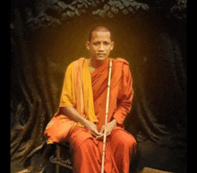 a monk in a red robe is sitting under a tree with a cane .