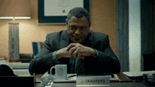 a man in a suit and tie sits at a desk with a name plate that says j crawford