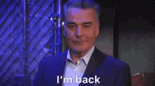 a man in a suit is standing in front of a chain link fence and says `` i 'm back '' .