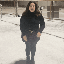 a woman standing in the snow wearing a black turtleneck and plaid skirt
