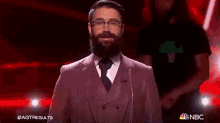 a man with a beard and glasses stands in front of a microphone with the nbc logo on the bottom right