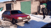 a red car is parked in the snow in front of a garage with a man walking by