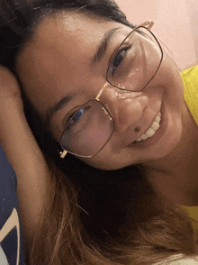 a woman wearing glasses and a yellow shirt smiles with her hand on her forehead