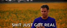a man in a blue shirt is standing in a field with the words `` shit just got real '' above him .