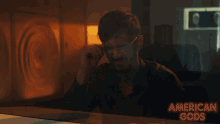 a man sits at a table with a sign that says american gods behind him