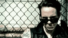 a man wearing sunglasses and a leather jacket is standing next to a chain link fence .