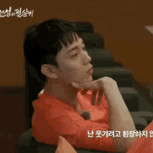 a man in a red shirt is sitting in a chair with his hand under his chin .