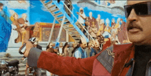 a man wearing sunglasses and a red jacket holds a gun in front of a staircase