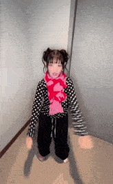 a little girl wearing a polka dot shirt and a pink scarf is standing in a hallway .