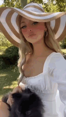 a woman wearing a white dress and a large hat is holding a black cat .