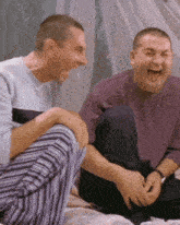 two men sit on a bed laughing together