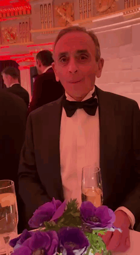a man in a tuxedo is sitting at a table with flowers and a glass of champagne .