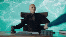 a bald man in a suit and tie is sitting at a desk with his arms outstretched