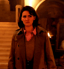 a woman wearing a trench coat and a pink shirt stands in a dark room