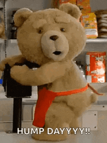 a teddy bear wearing a red apron is hugging a machine and says hump day yy !