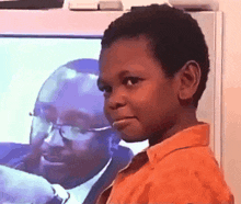 a young boy in an orange shirt looks at the camera in front of a tv screen